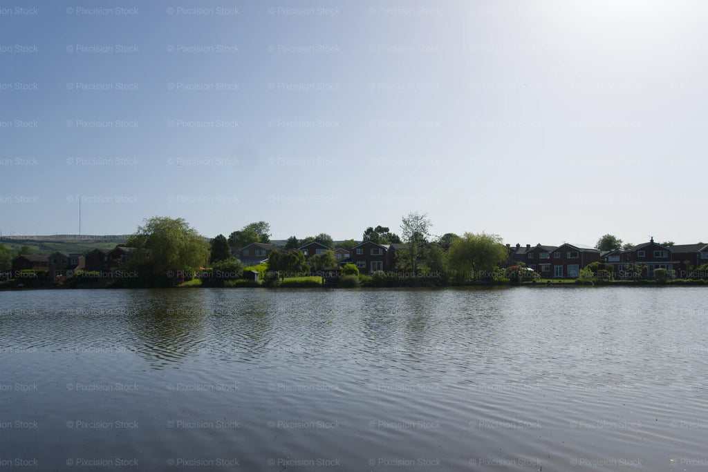 A wide, scenic shot of a calm lake with gentle ripples in the foreground, bordered by a row of suburban residential houses and lush green trees under a bright, clear blue sky.