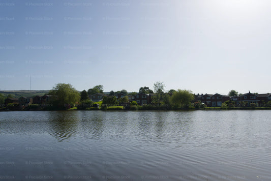 A wide, scenic shot of a calm lake with gentle ripples in the foreground, bordered by a row of suburban residential houses and lush green trees under a bright, clear blue sky.