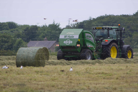 Green tractor baling hay in a field with trees in the background