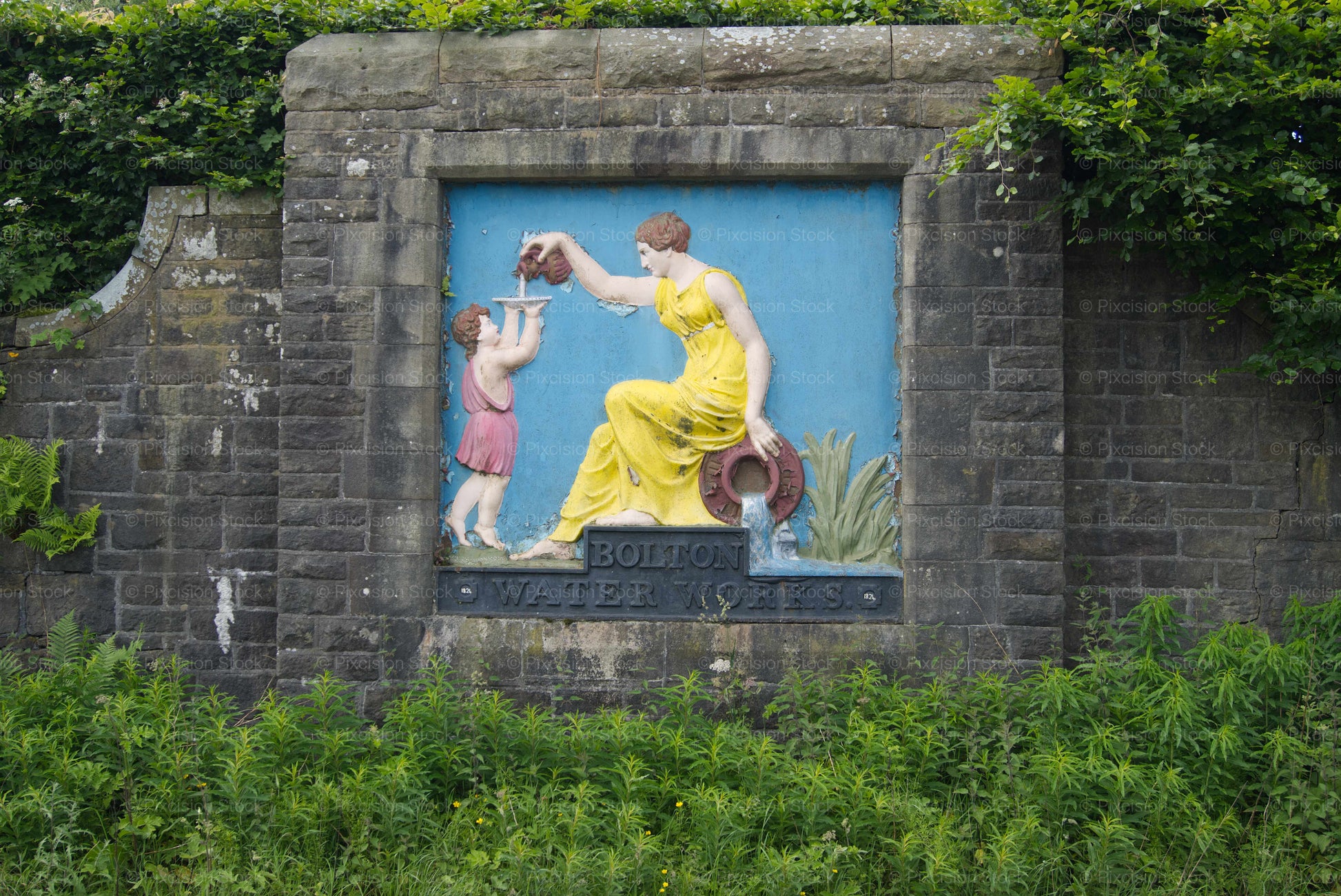 A close-up view of the Grade II listed Bolton Water Works stone relief plaque embedded in a dark stone wall on Belmont Road. The painted scene shows an allegorical female figure and child in vibrant colors above a cast-iron nameplate, surrounded by natural green foliage.