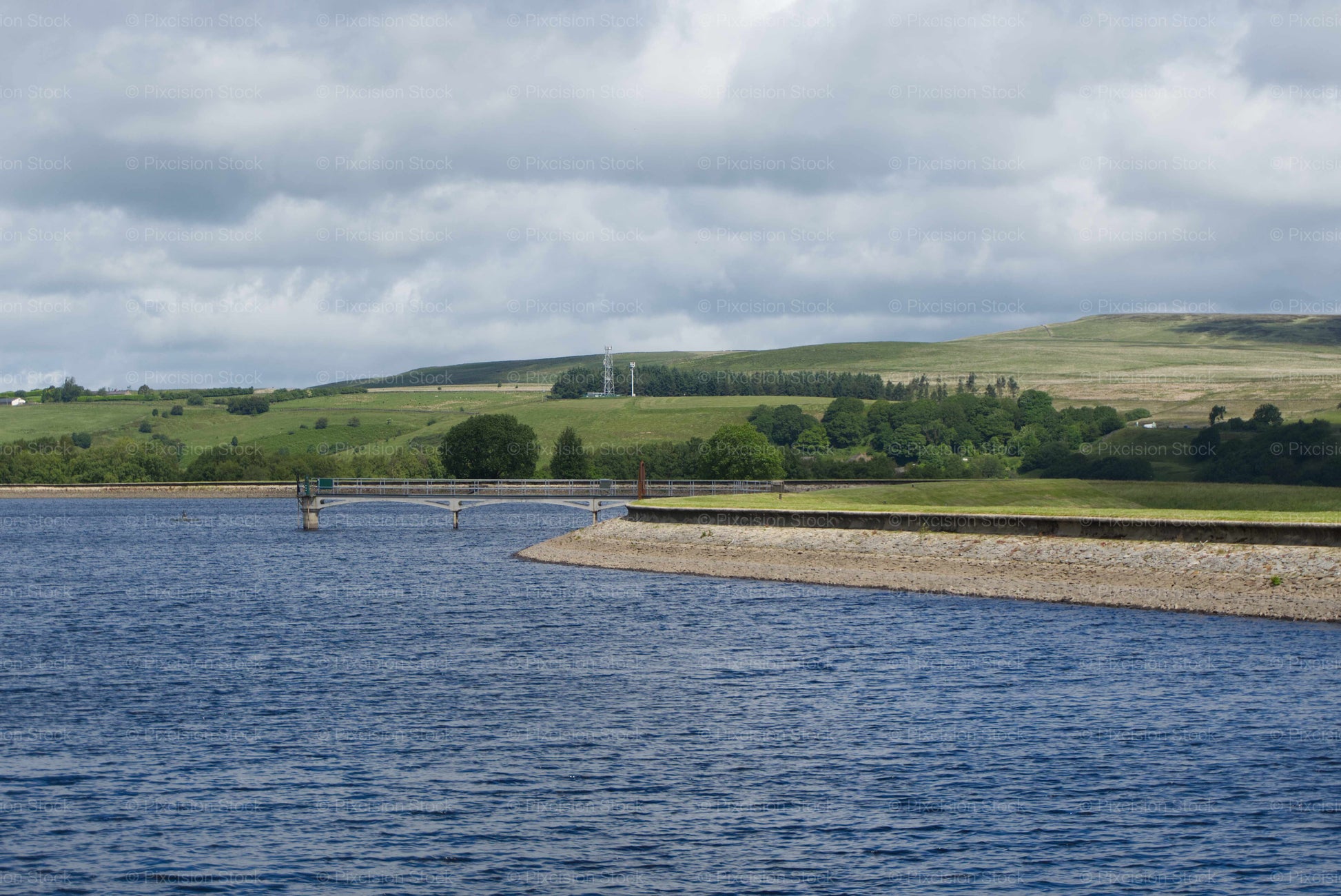 A scenic landscape view looking across the deep blue waters of Springs Reservoir. The shot features the stone embankment, a concrete gantry walkway bridge, an outlet valve tower, and the rolling green pastures of the West Pennine Moors stretching out under a cloudy sky.