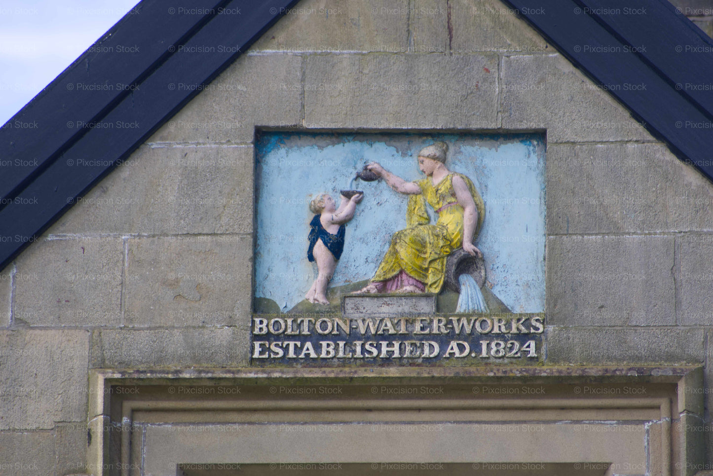 A close-up view of the historic bas-relief plaque set into the stone gable of Waterworks Cottage. The artwork features a woman in a yellow gown pouring water into a bowl held by a child against a blue background, above the inscription Bolton Waterworks Established A.D. 1824.