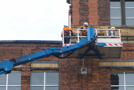 Two workers on a cherry picker platform against a brick building.
