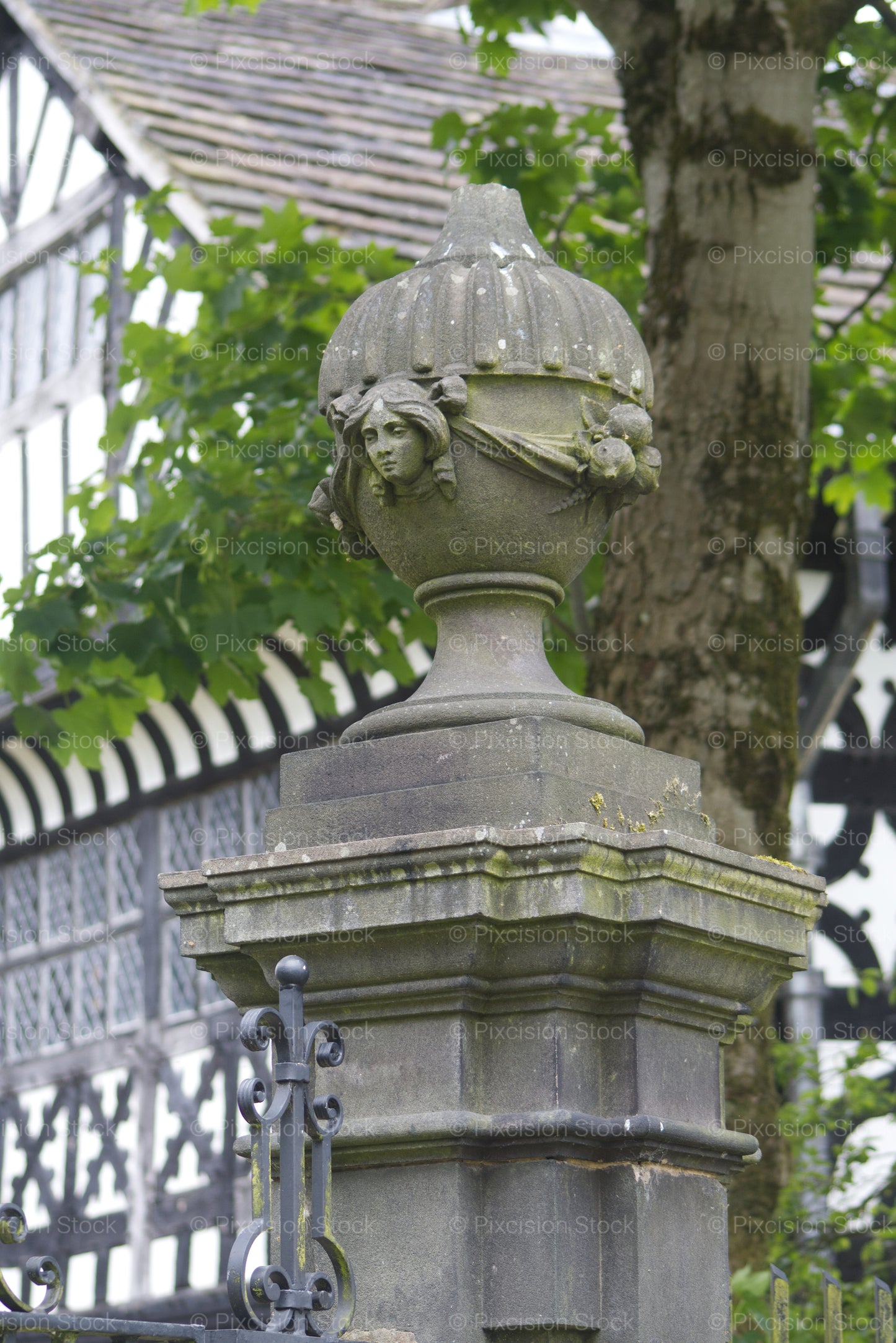 An ornate stone urn finial with a carved face atop a pillar, set against a historic half-timbered Tudor building.