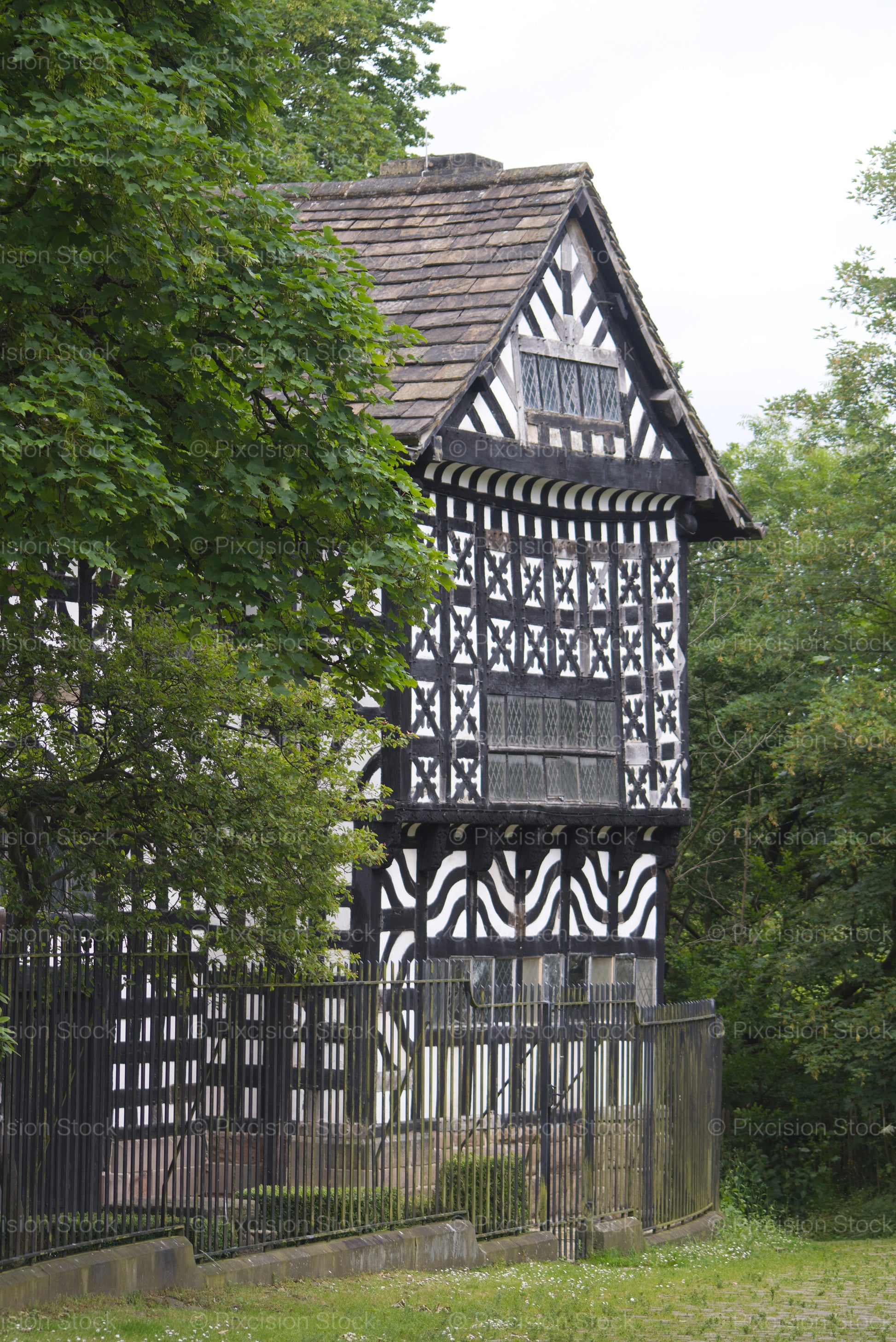 A historic black and white half-timbered building partially obscured by green trees behind a metal fence.