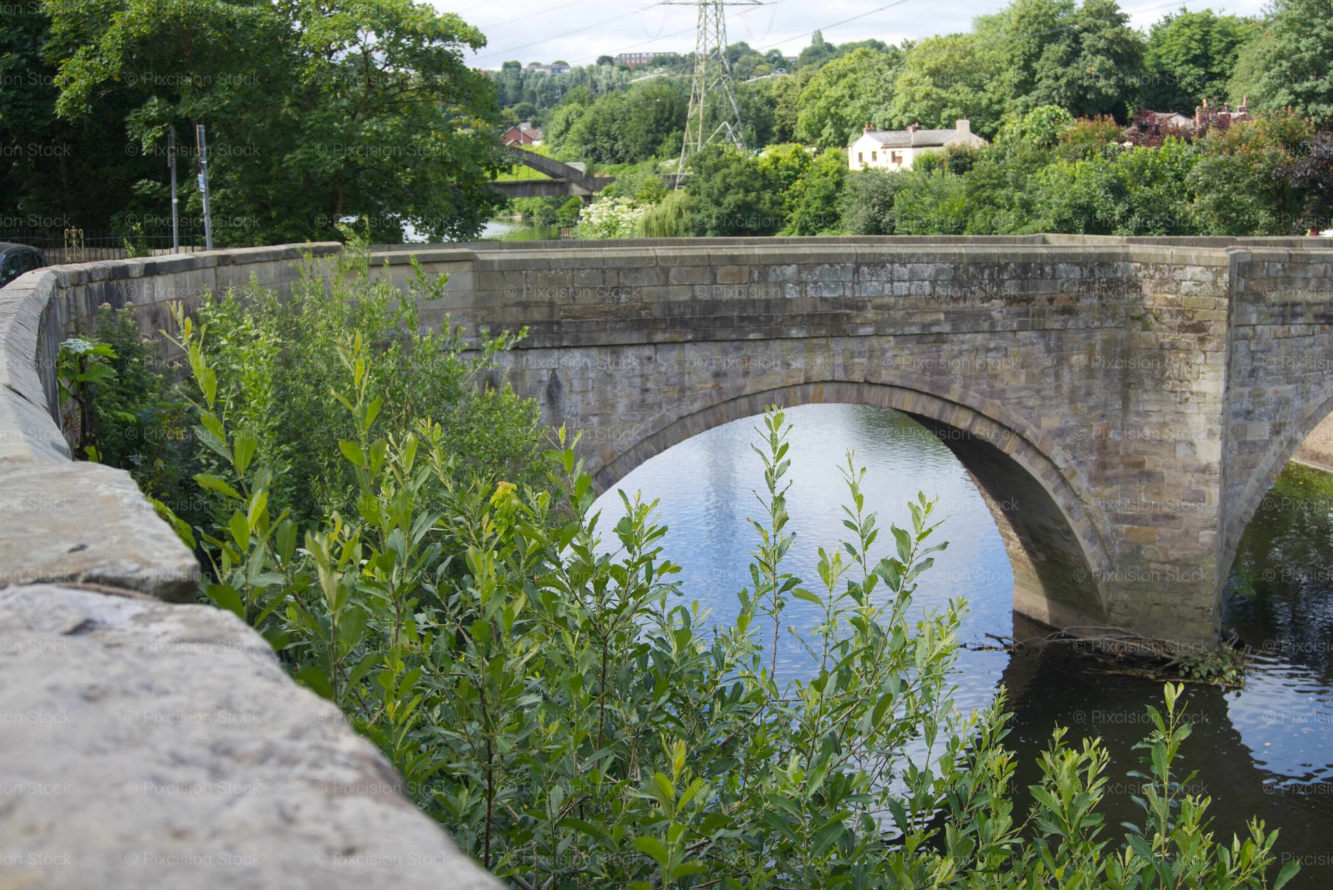 The historic 1677 stone arches of Ringley Old Bridge crossing over the calm River Irwell, framed by lush green bushes in the foreground.