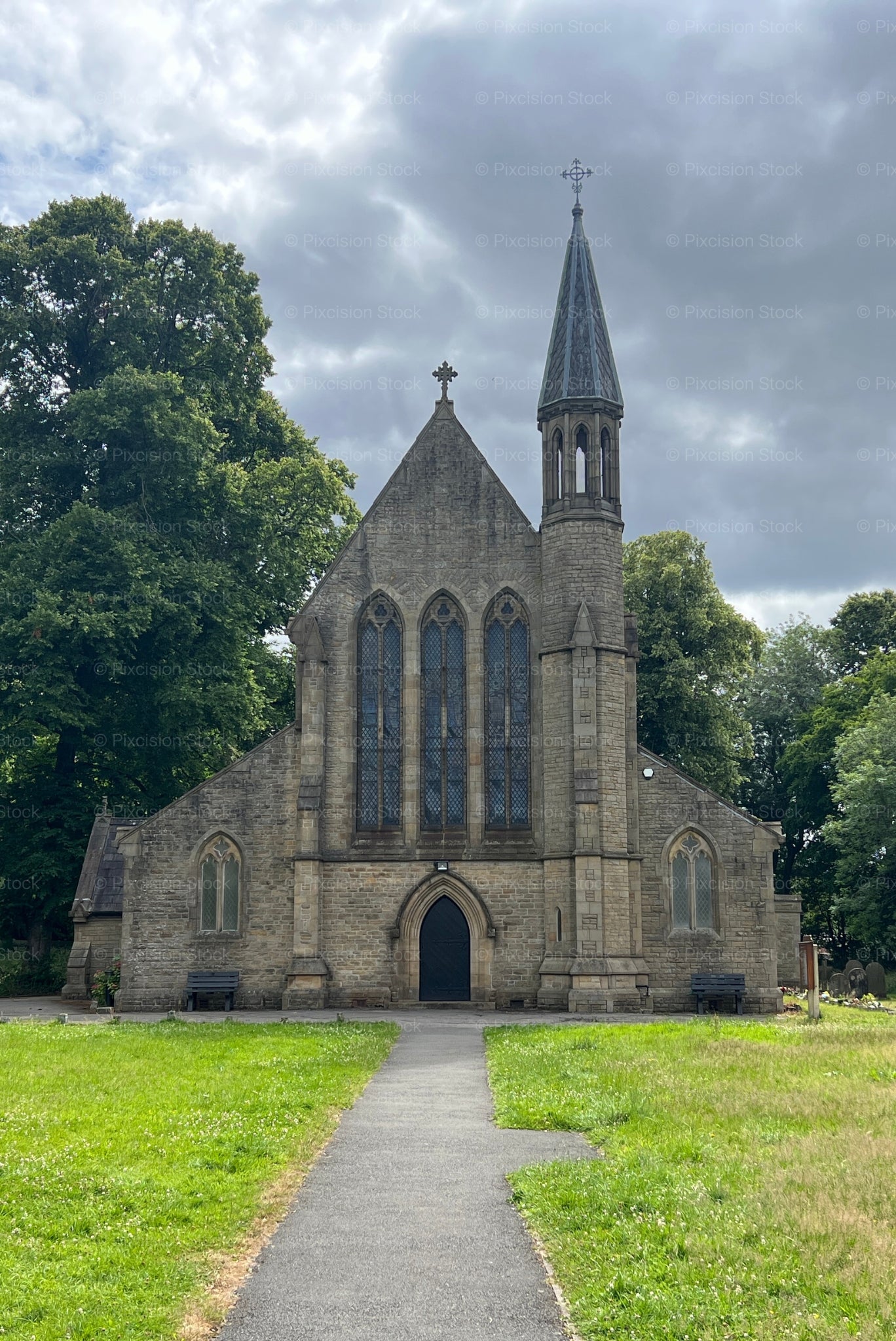 A centered front view of the stone St Saviour's Church in Radcliffe, England, showing a path leading through a grassy churchyard to the entrance, surrounded by dense trees.