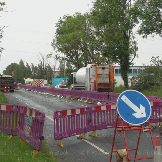 A video of a road construction zone with purple barriers, temporary traffic lights, and directional signs as a large lorry drives away down a narrow road.