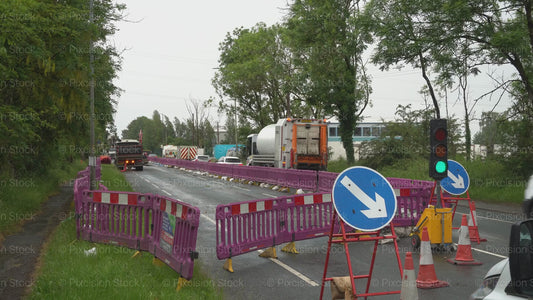 A video of a road construction zone with purple barriers, temporary traffic lights, and directional signs as a large lorry drives away down a narrow road.