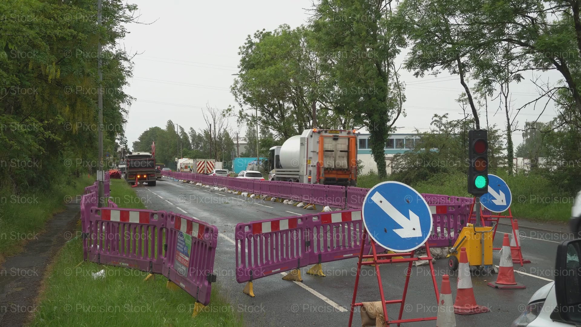 A video of a road construction zone with purple barriers, temporary traffic lights, and directional signs as a large lorry drives away down a narrow road.