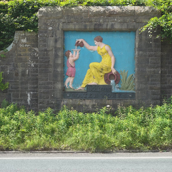 A steady video frame showing the historic, Grade II listed Bolton Water Works painted relief plaque embedded into a roadside stone wall on Belmont Road, surrounded by thick green hedges and roadside foliage.