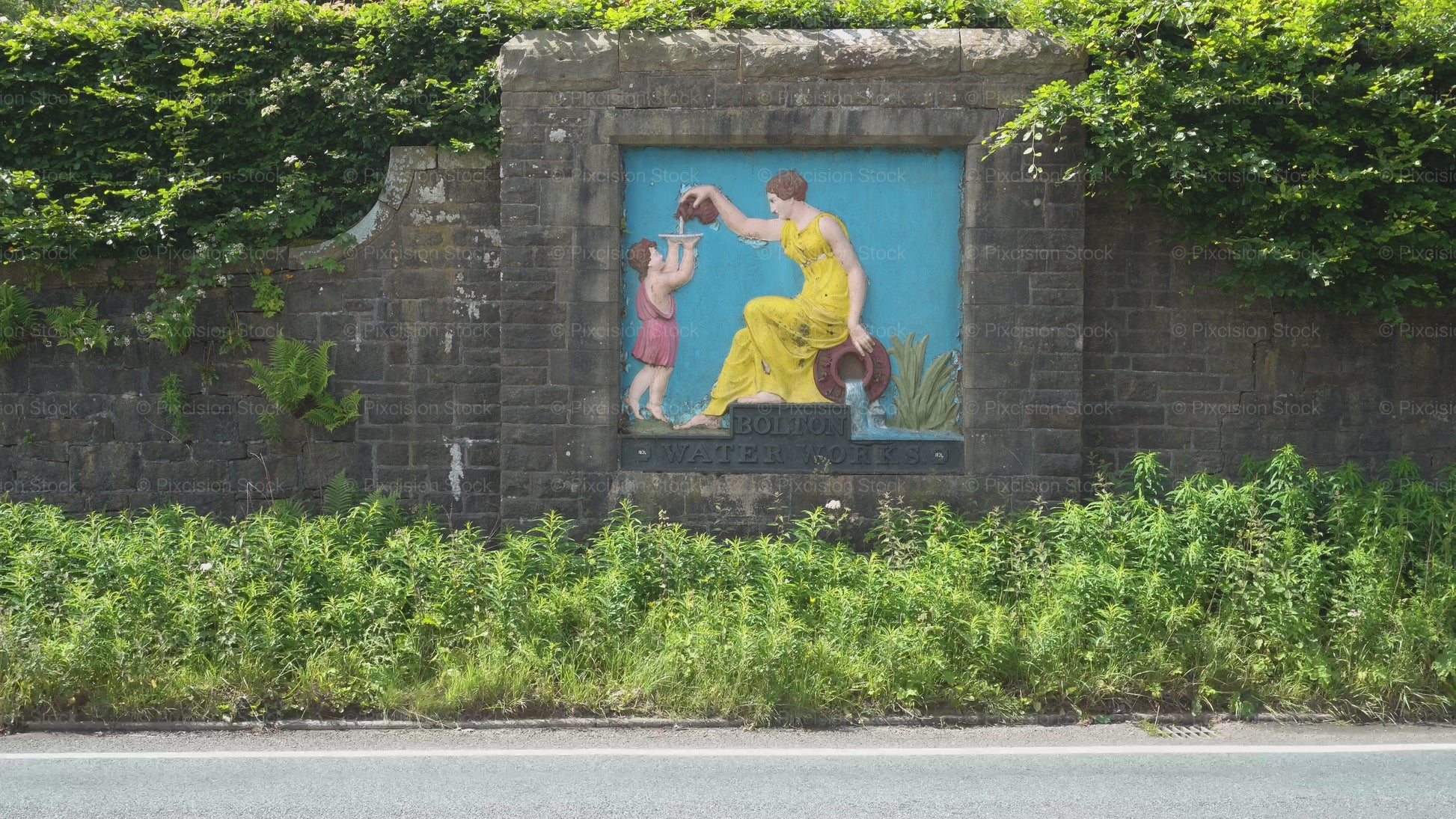 A steady video frame showing the historic, Grade II listed Bolton Water Works painted relief plaque embedded into a roadside stone wall on Belmont Road, surrounded by thick green hedges and roadside foliage.