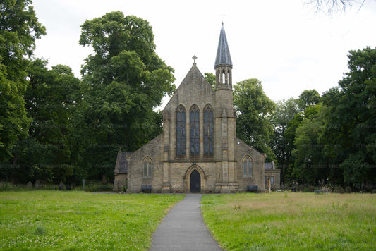 A centered front view of the stone St Saviour's Church in Radcliffe, England, showing a path leading through a grassy churchyard to the entrance, surrounded by dense trees.